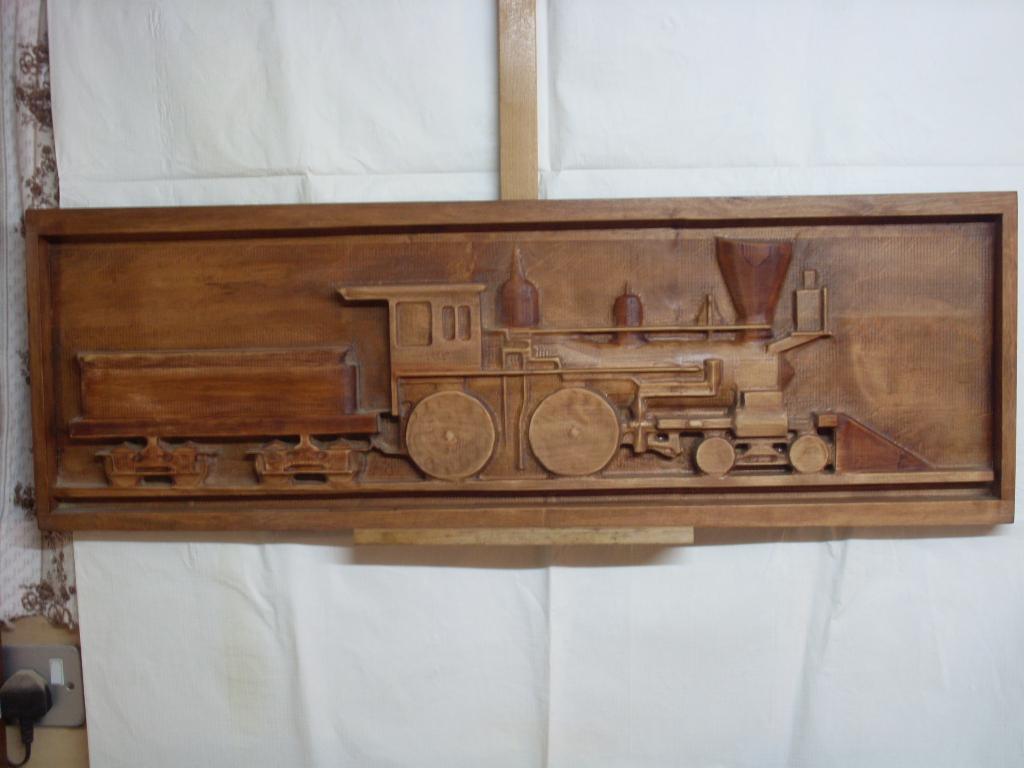 Drewniana płaskorzeźba przedstawiająca lokomotywę z wagonem, umieszczona w prostokątnej ramie, zawieszona na białym tle.