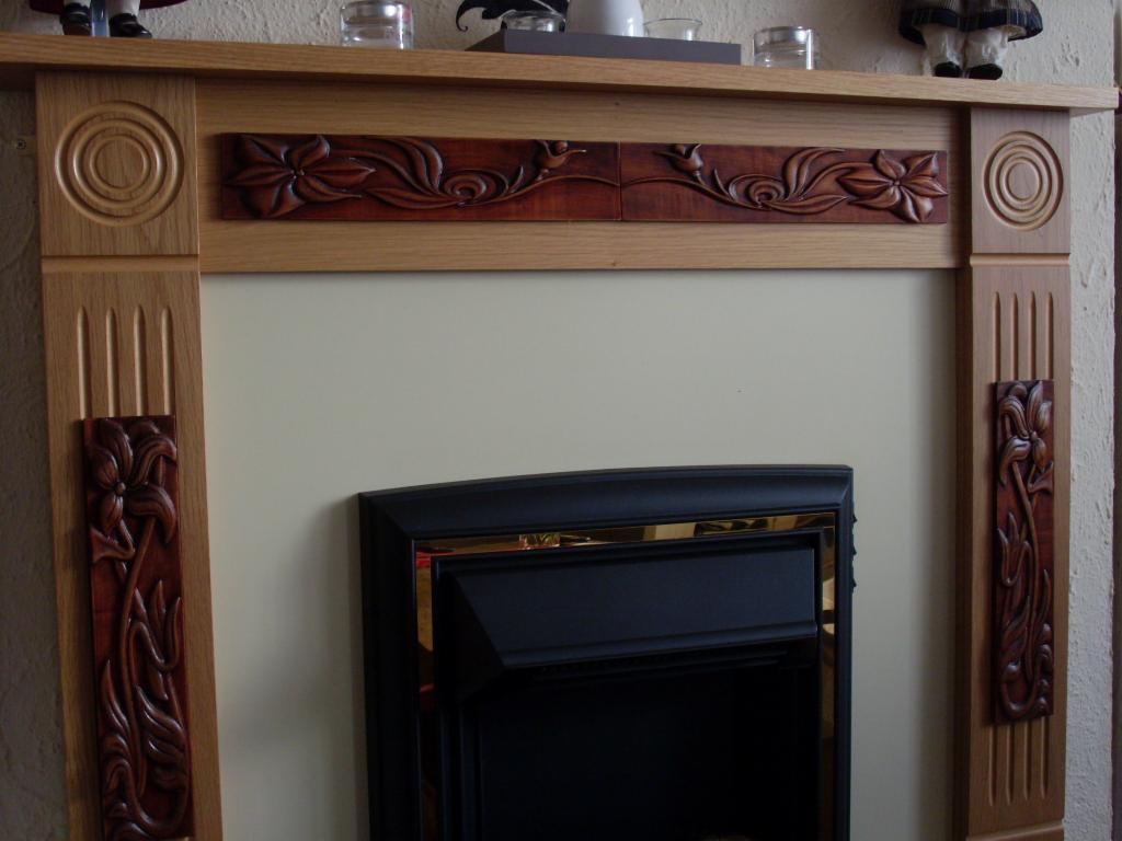 Ozdobny kominek z drewnianą obudową z rzeźbionymi kwiatowymi motywami i czarnym wkładem kominkowym z elementami w kolorze złota.