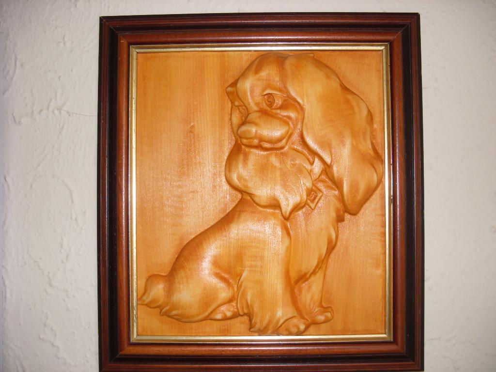 Drewniana płaskorzeźba przedstawiająca psa rasy spaniel w brązowej ramie z elementami złota, umieszczona na kremowej ścianie.