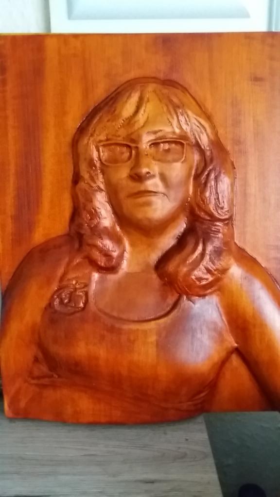 Ręcznie rzeźbiony w drewnie portret kobiety w okularach, z widocznym detalem przypinki na koszulce, na jasnym tle.