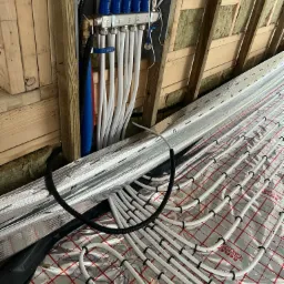 Rozdzielacz z zaworami Efar i rurami podłogowymi Reflex Termosystems na folii refleksyjnej z czerwoną kratką, widok na drewnianą konstrukcję dachu z izolacją.