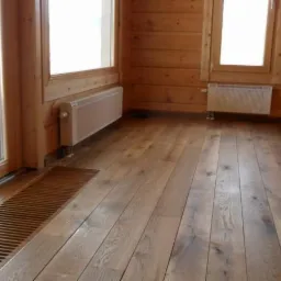 Jasne, drewniane wnętrze z podłogą z desek, grzejnikami ściennymi i kratką podłogową, widok na okno z drewnianą ramą.