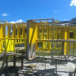 Metalowa konstrukcja pomalowana na jaskrawy żółty kolor, wsparta na prowizorycznych rusztowaniach na zewnątrz, w tle błękitne niebo i drzewa.