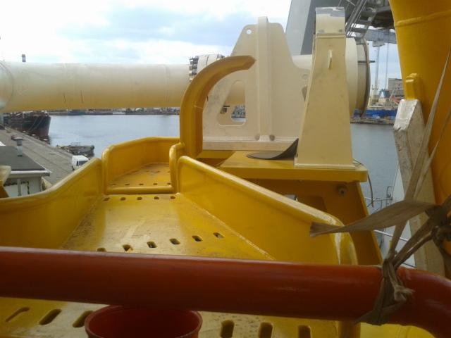 Żółta konstrukcja metalowa z elementami pomalowanymi na biało, widoczna na tle portu z zacumowanymi statkami i elementami dźwigu.