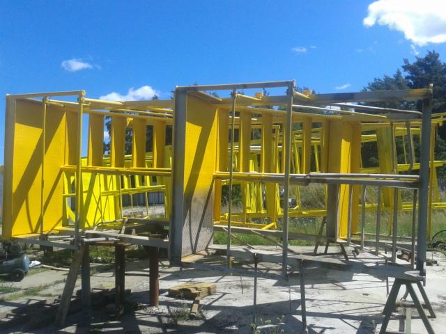 Metalowa konstrukcja pomalowana na jaskrawy żółty kolor, wsparta na prowizorycznych rusztowaniach na zewnątrz, w tle błękitne niebo i drzewa.
