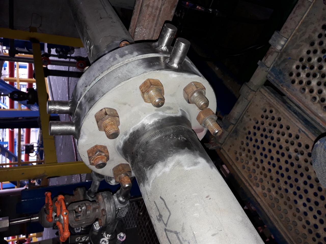 Metalowa rura z kołnierzem i śladami spawania, zardzewiałe śruby mocujące, w tle elementy konstrukcji przemysłowej