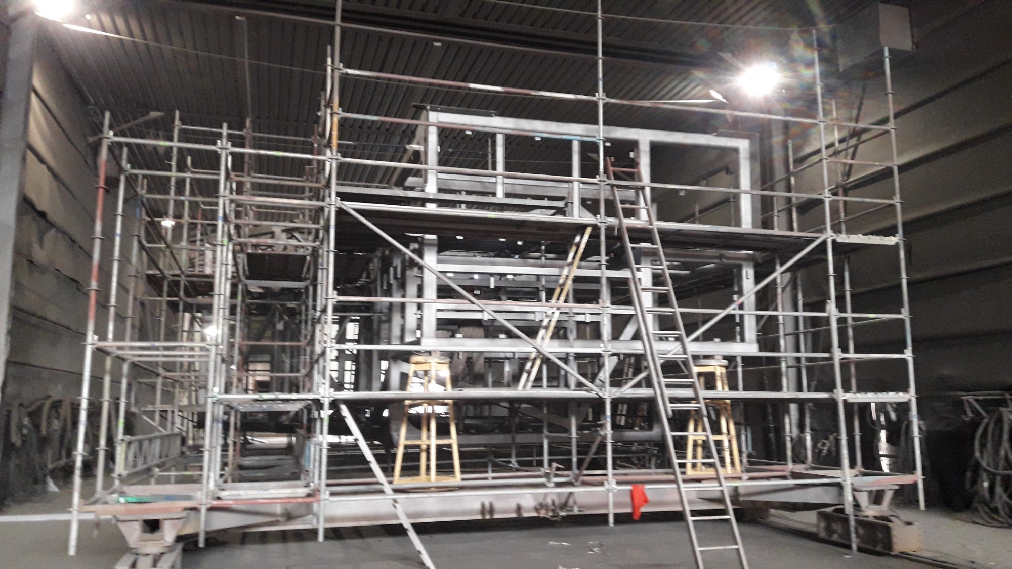 Konstrukcja metalowa w hali produkcyjnej, otoczona rusztowaniem i schodami, oświetlona jasnym światłem