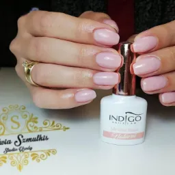 Delikatny, pudrowo-różowy manicure hybrydowy na krótkich, owalnych paznokciach, prezentowany obok buteleczki bazy Indigo Natural. Na palcu serdecznym złoty pierścionek z oczkiem, w tle logo salonu.