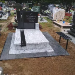 Nowoczesny nagrobek z czarnego i szarego granitu na cmentarzu, z wygrawerowanym krzyżem i inskrypcjami, otoczony brukowaną ścieżką i ziemią, widoczne inne nagrobki w tle.