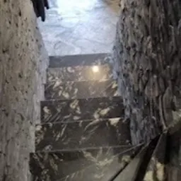 Wąskie schody wewnętrzne wykończone czarnym kamieniem z białymi żyłkami, otoczone ścianami z szarego kamienia łupanego, widok z góry na stopnie i fragment butów osoby robiącej zdjęcie.