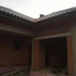 Surowy stan budynku z częściowo ukończonym dachem z ciemnej dach&oacute;wki. Widoczne ceglane ściany, betonowe nadproża i pozostałości materiał&oacute;w budowlanych.