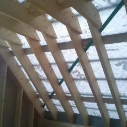 Wnętrze domu w trakcie budowy: widok na drewnianą konstrukcję dachu z elementami izolacji i zielonymi wzmocnieniami, oświetlone naturalnym światłem.