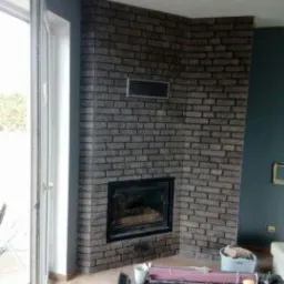 Narożny kominek obłożony szarą cegłą klinkierową, z widocznym wkładem kominkowym i fragmentem ściany pomalowanej na ciemny, grafitowy kolor. W tle widoczne otwarte drzwi balkonowe i fragment mebli.