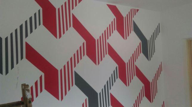 Ściana z geometrycznym wzorem w kolorach czerwonym i grafitowym, wykonanym techniką malarską, z fragmentem drabiny malarskiej w lewym dolnym rogu.