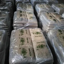 Stosy worków z granulatem opałowym z widocznym nadrukiem 'Wood Pellet' i motywem drzew iglastych, zapakowane w folię, magazynowane w hali.