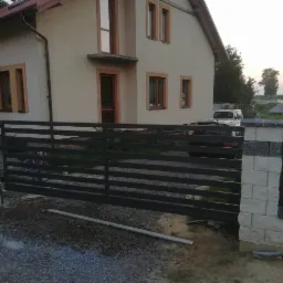 Czarna, pozioma brama wjazdowa z paneli stalowych, osadzona między słupkami z szarej kostki betonowej, na tle domu jednorodzinnego i zaparkowanego samochodu.