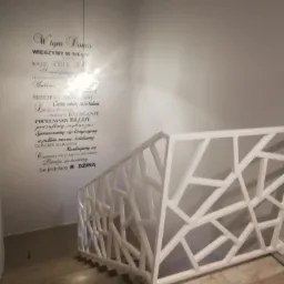 Biała balustrada wewnętrzna o geometrycznym wzorze na schodach, z dekoracyjnym napisem na ścianie w tle i wiszącą lampą.