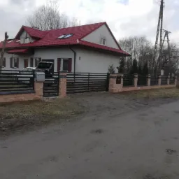 Nowoczesne ogrodzenie posesji z czarnymi, poziomymi przęsłami i słupkami z imitacji kamienia, widoczne na tle domu z czerwoną dachówką i szarym niebem.