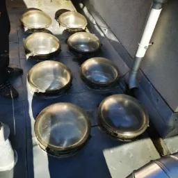 Szklane, okrągłe świetliki dachowe na płaskim dachu, z widoczną rynną spustową i fragmentem instalacji wentylacyjnej, w słoneczny dzień.