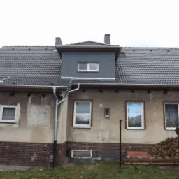 Dom z nowym, ciemnoszarym dachem z dachówki ceramicznej, widoczne rynny, komin i okno dachowe w szarym lukarnie, elewacja częściowo z cegły, stos drewna na podwórku.