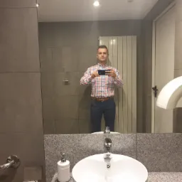 Mężczyzna robi selfie w łazience z szarymi płytkami i białą umywalką. Widoczne dozowniki i suszarki do rąk. Ujęcie z odbicia w lustrze.