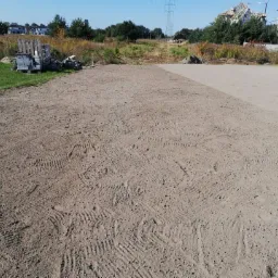 Przygotowany teren pod trawnik z rolki, widoczne ślady po maszynach, w tle paleta z kostką brukową i budowa domu.