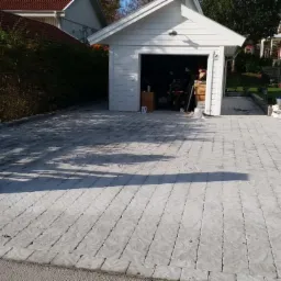 Ułożona szara kostka brukowa na podjeździe przed garażem, widoczny fragment ogrodzenia i pracownik w tle.