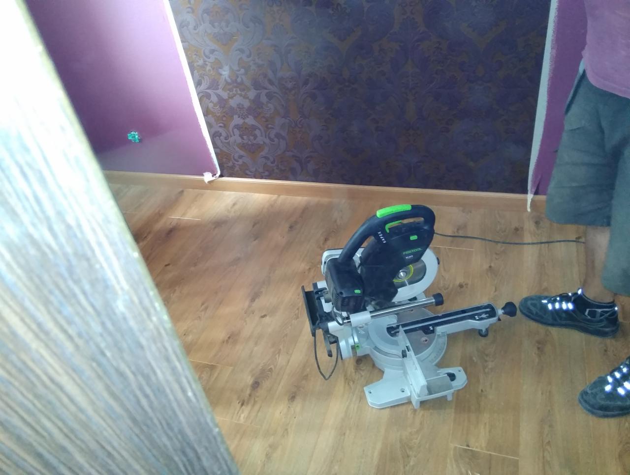 Ukośnica Festool KAPEX KS 60 na podłodze z paneli podczas montażu listew przypodłogowych, widoczny fragment ściany z tapetą w tle.