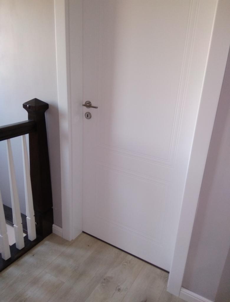 Zamknięte, białe drzwi wewnętrzne z srebrną klamką, widoczne od strony schodów z drewnianą balustradą.