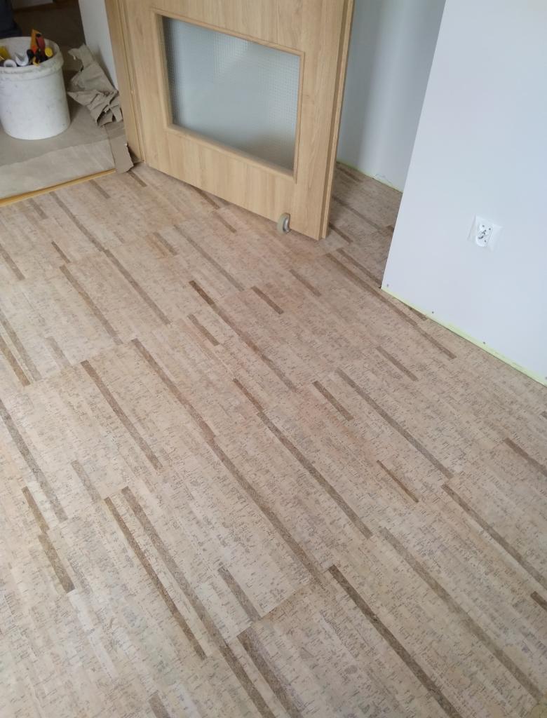 Układanie paneli korkowych na podłodze w pokoju, widoczna drewniana ościeżnica drzwi z szybą i biała ściana z gniazdkiem elektrycznym.