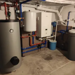 Zestaw urządzeń w kotłowni: szary zasobnik ciepłej wody z manometrem, biały bojler, biała jednostka centralna z wyświetlaczem i szary zmiękczacz wody, połączone rurami w kolorach czerwonym...