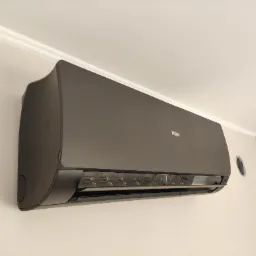 Czarny klimatyzator Haier zamontowany na jasnej ścianie pod sufitem z ozdobnym gzymsem. Widoczny okrągły otwór wentylacyjny w ścianie obok.