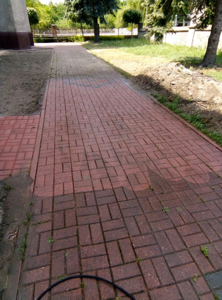 Częściowo umyta, czerwona kostka brukowa ułożona w jodełkę, widoczne mokre i suche fragmenty, trawa rosnąca między kostkami, obok trawnik.