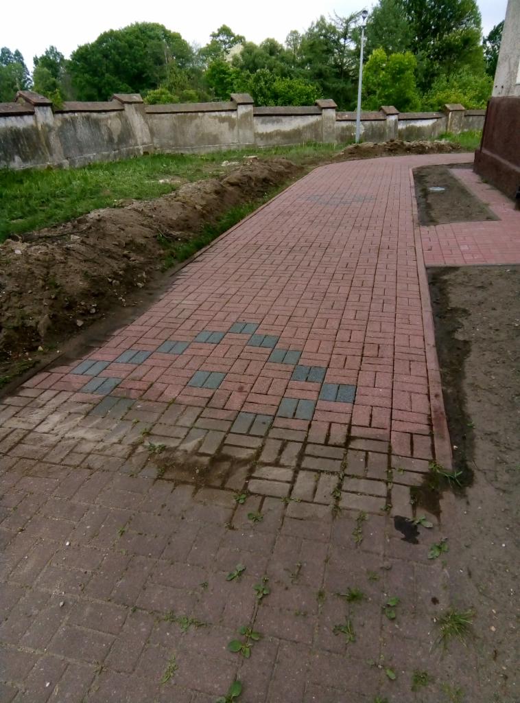 Ułożona ścieżka z czerwonej kostki brukowej z elementami dekoracyjnymi z szarej kostki, widoczne ślady po budowie i fragment trawnika z ziemią.