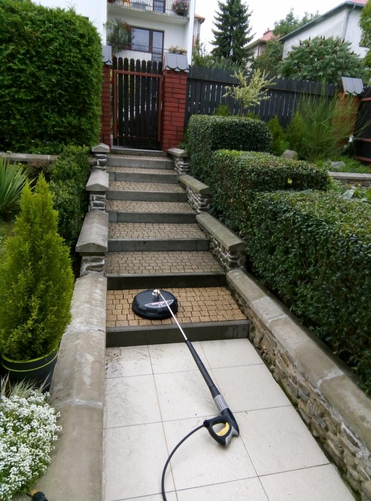 Czyszczenie kamiennych schodów prowadzących do ogrodu za pomocą myjki ciśnieniowej z okrągłą szczotką. Widoczne rośliny ozdobne i elementy architektury ogrodowej.