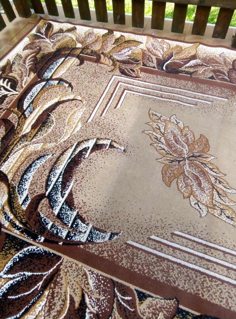 Beżowy dywan z brązowo-białym roślinnym wzorem, leżący na balkonie z drewnianą balustradą, widok z góry.