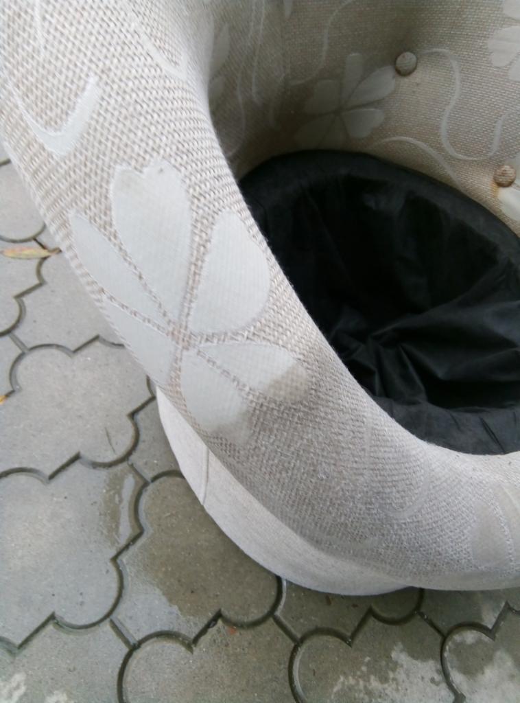 Fragment fotela obrotowego z beżową tapicerką w kwiatowy wzór, z widocznym wnętrzem wyłożonym czarnym materiałem, stojącego na mokrej, szarej kostce brukowej.