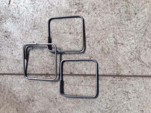 Trzy kwadratowe, metalowe strzemiona z żebrowanej stali ułożone na szarym, betonowym podłożu z widocznymi rysami i zabrudzeniami.