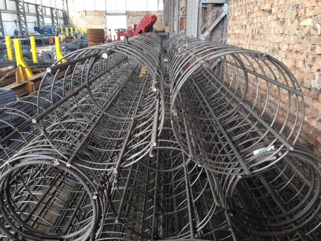 Klatki zbrojeniowe o okrągłym przekroju składowane w hali produkcyjnej, widoczny surowy mur z cegły w tle.