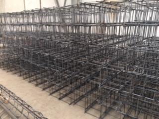 Stosy gotowych, przestrzennych szkieletów zbrojeniowych ułożonych jeden na drugim w hali magazynowej, widoczne spawane łączenia prętów.
