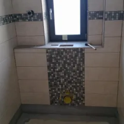 Montaż beżowych płytek z dekoracyjną mozaiką w łazience, widoczne żółte rury odpływowe i surowa podłoga w trakcie remontu.