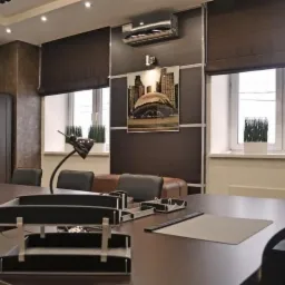 Wnętrze biura z dużym drewnianym stołem, lampką biurkową w stylu retro, obrazem panoramy miasta na ścianie i brązowymi roletami rzymskimi w oknach.