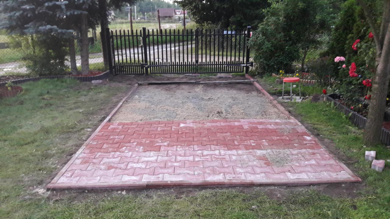 Częściowo ułożona nawierzchnia z czerwonej kostki brukowej na przygotowanym podłożu z piasku, widok z perspektywy trawnika w ogrodzie, brama w tle.