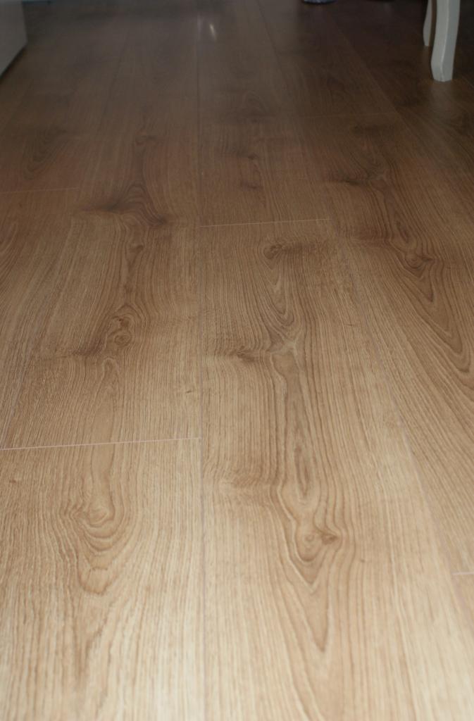 Jasne panele podłogowe z wyraźnym wzorem drewna, ułożone wzdłuż pomieszczenia, z fragmentem białej nogi mebla w prawym górnym rogu.