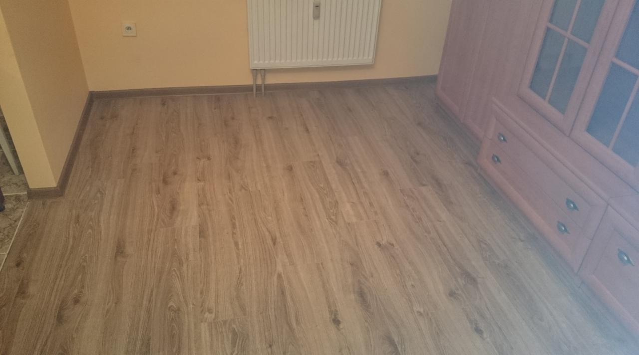 Ułożone panele podłogowe w pokoju z beżowymi ścianami, widoczny fragment szafy i grzejnik.
