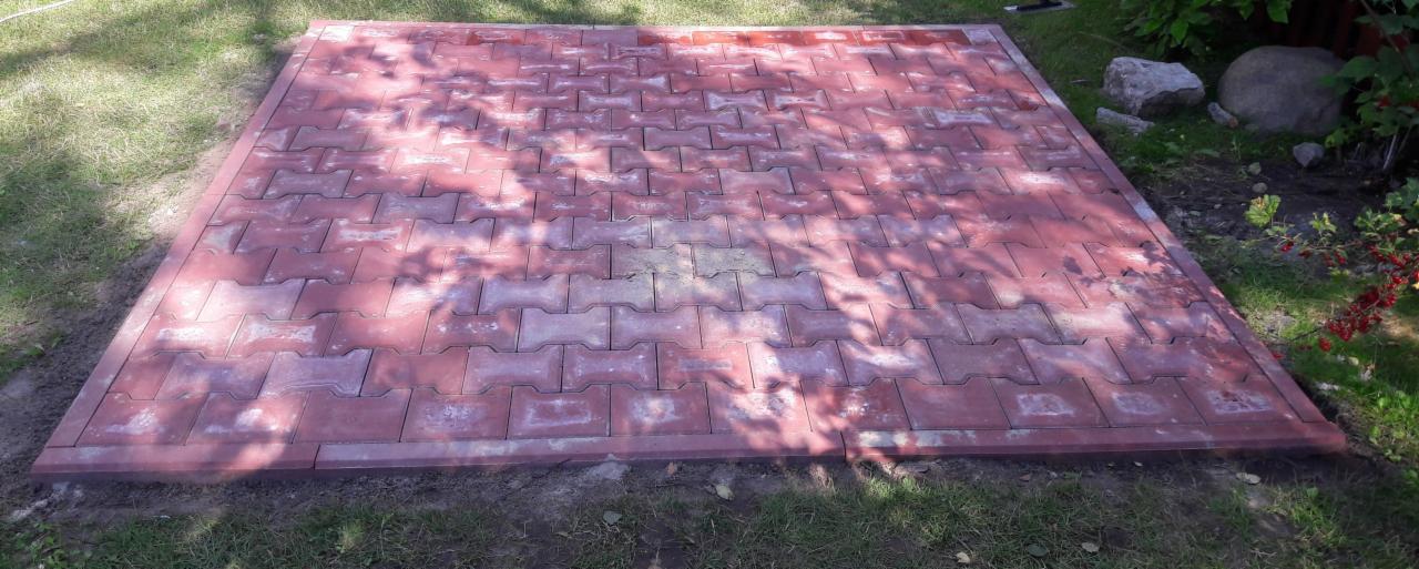 Ułożona, czerwona kostka brukowa na trawniku, widoczne ślady piasku między kostkami, w tle roślinność ogrodowa.