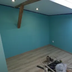 Pok&oacute;j po malowaniu ścian na turkusowo, z drewnianą belką stropową, oświetleniem punktowym i przygotowanymi listwami przypodłogowymi oraz narzędziami na podłodze z paneli.