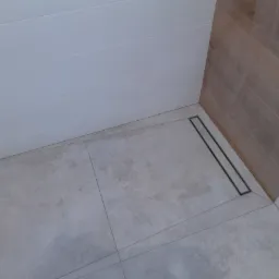 Nowoczesny odpływ liniowy w kabinie prysznicowej wyłożonej szarymi płytkami podłogowymi i białymi płytkami ściennymi z elementami dekoracyjnymi imitującymi drewno.