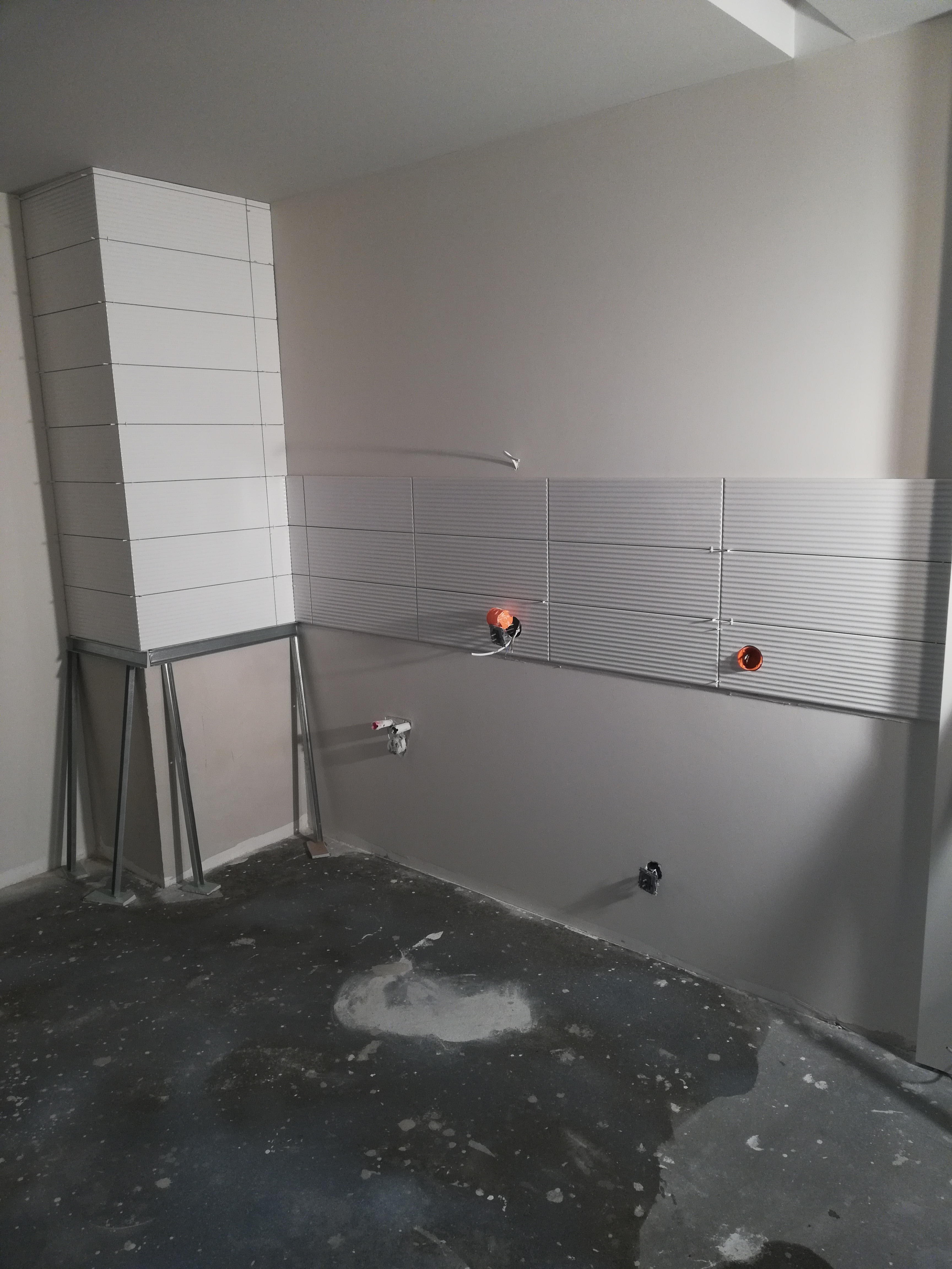 Świeżo położone, poziome, szare płytki z delikatnymi rowkami na ścianie łazienki, częściowo zamontowane na metalowej konstrukcji, z widocznymi otworami na instalacje i niedokończoną podłogą.