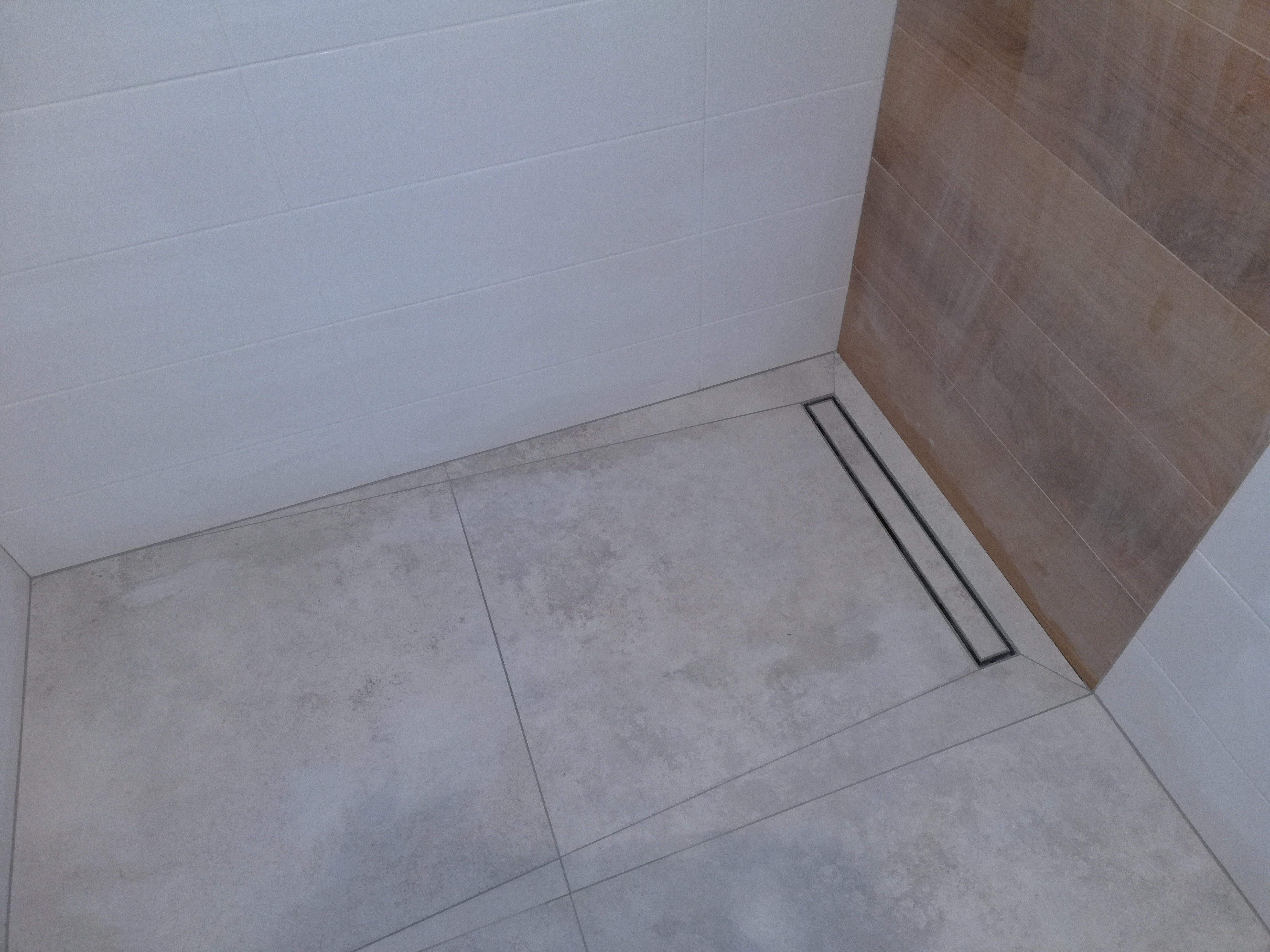 Nowoczesny odpływ liniowy w kabinie prysznicowej wyłożonej szarymi płytkami podłogowymi i białymi płytkami ściennymi z elementami dekoracyjnymi imitującymi drewno.